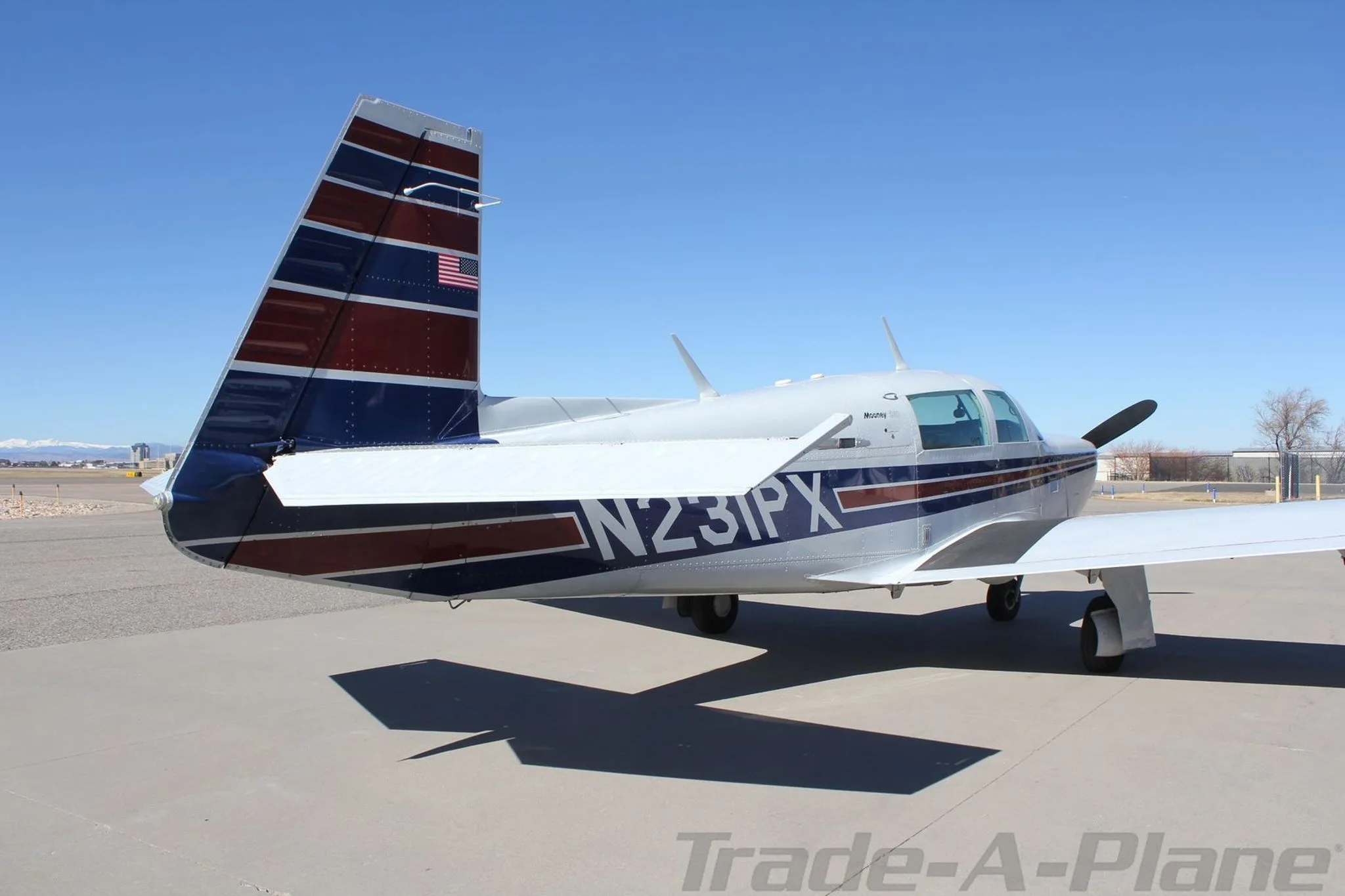 Rear-side view of a silver single-engine aircraft with blue and red striped tail and fuselage, parked on an airfield.