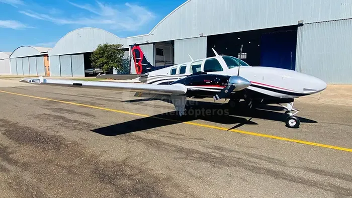 A white and black twin-engine piston aircraft with red accents parked on a tarmac in front of hangars under a blue sky.