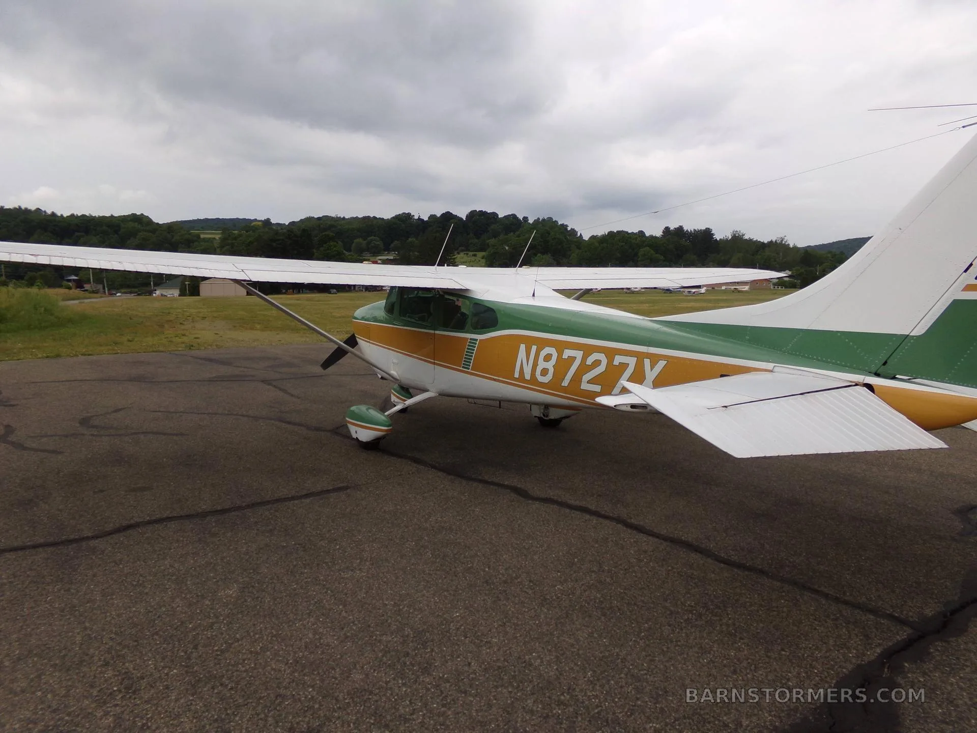 A green, gold, and white Cessna 172 aircraft with registration N8727X, viewed from the rear-left on an asphalt ramp under a cloudy sky.