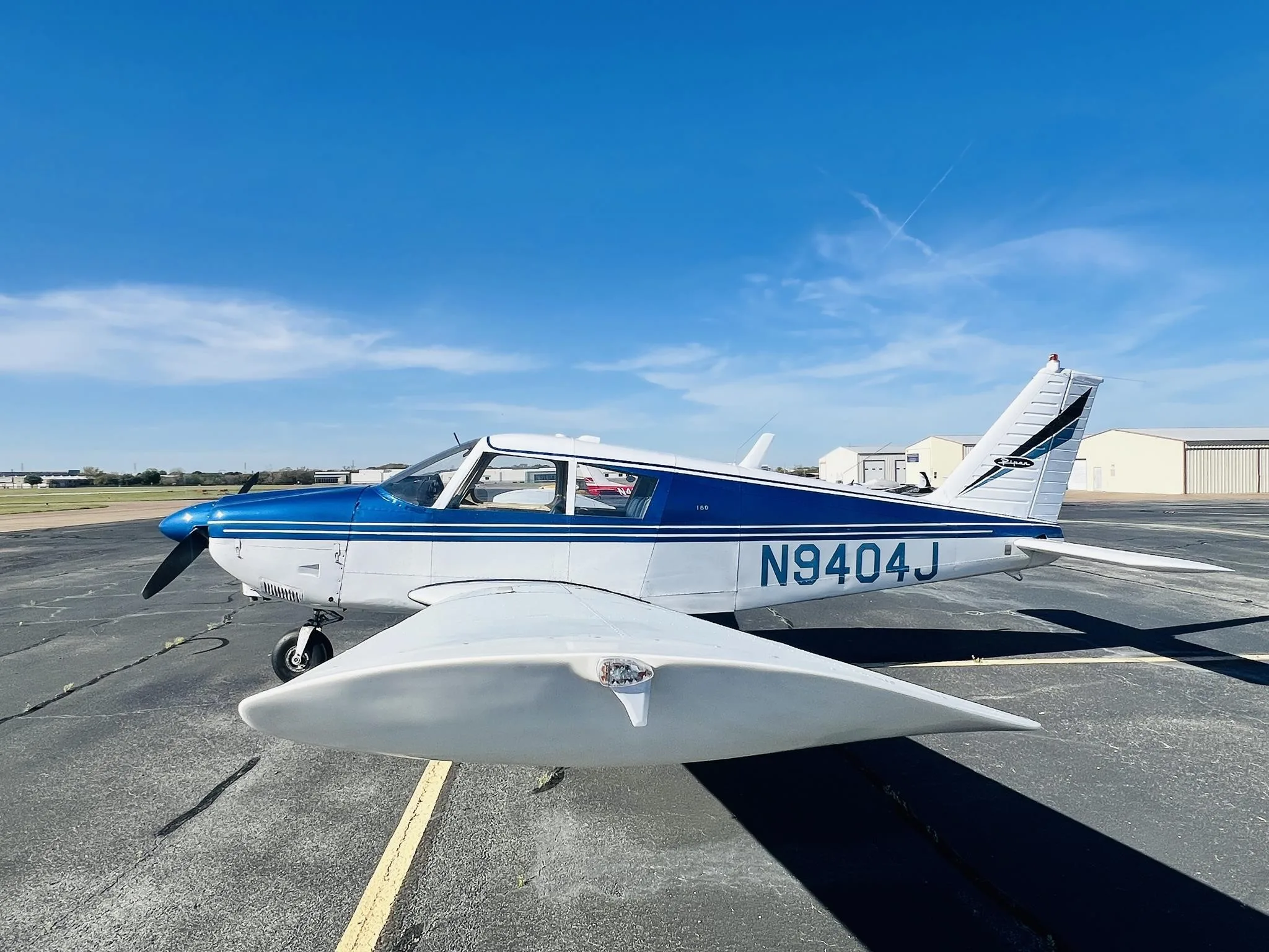 A white and blue single-engine propeller aircraft, tail number N9404J, is parked on an asphalt tarmac under a blue sky.