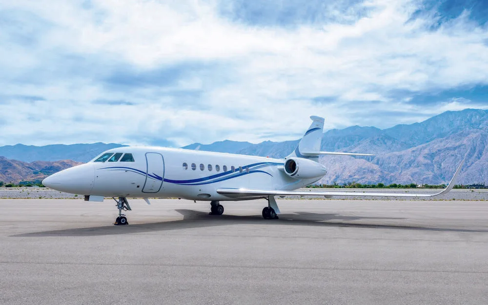A white Dassault Falcon 2000LX business jet with blue stripes on the tarmac, with mountains in the background.