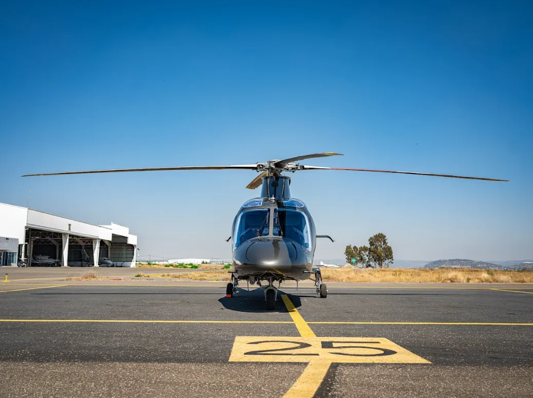 A grey AgustaWestland AW109 GrandNew helicopter is seen from the front, parked on an airfield with "25" runway markings.