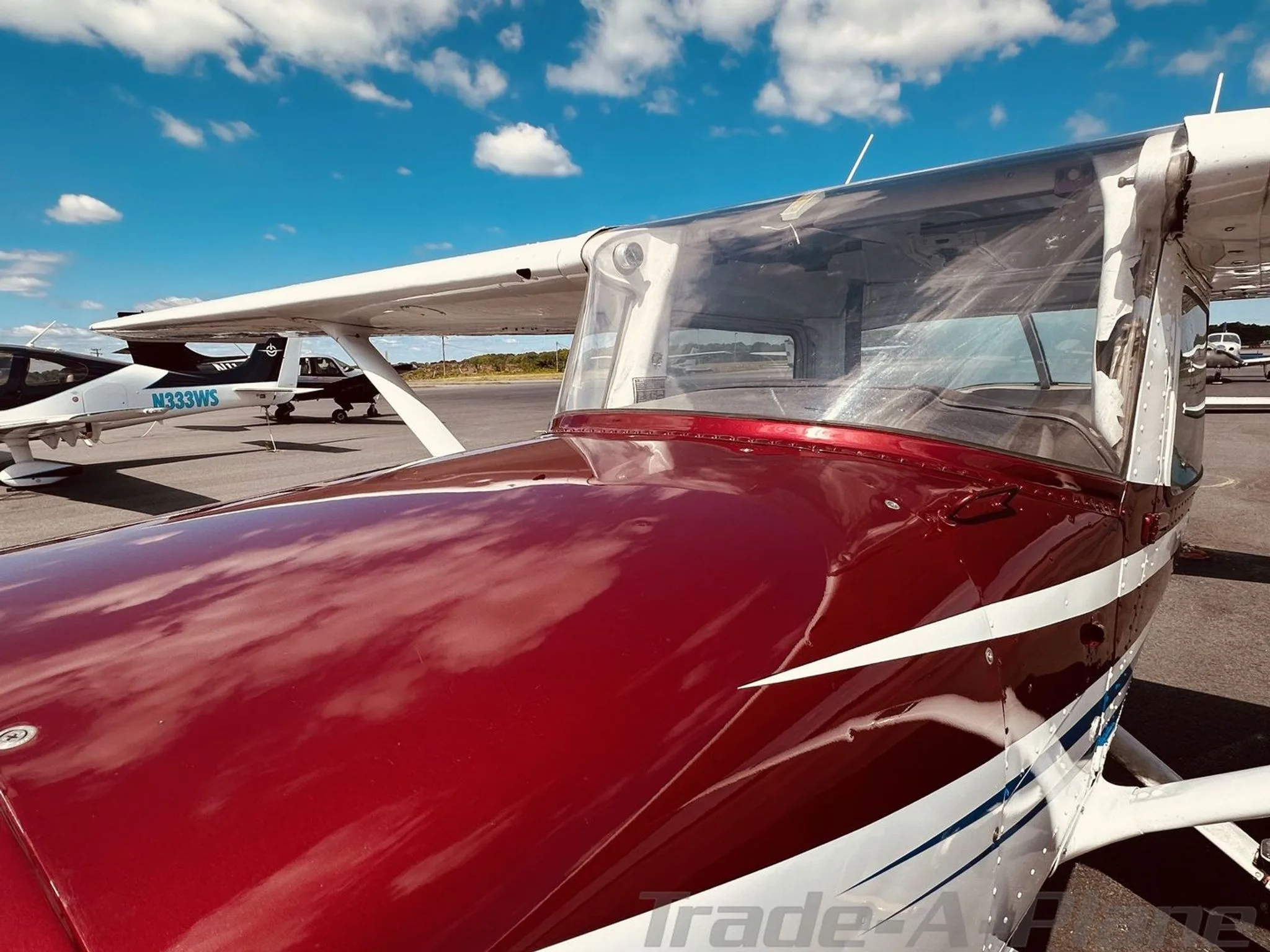A close-up view of the front fuselage and cockpit of a white Cessna 172 with maroon and blue stripes, parked on an airport tarmac.
