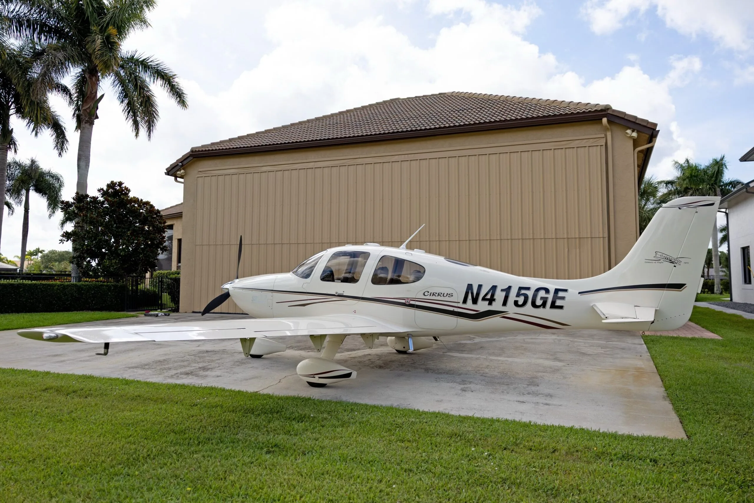 A white Cirrus SR22 aircraft with black and maroon stripes, registration N415GE, parked on a concrete pad next to a tan building.