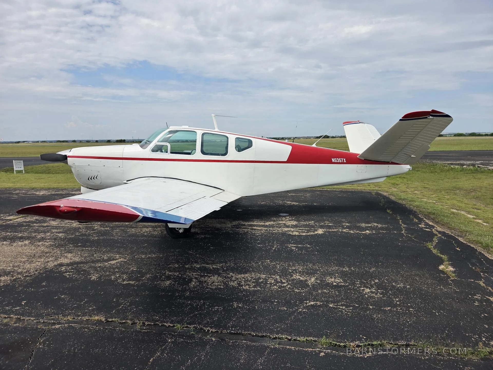 A white and red Beechcraft Bonanza aircraft with registration N535TX is parked on an asphalt ramp under a cloudy sky, viewed from the left side.