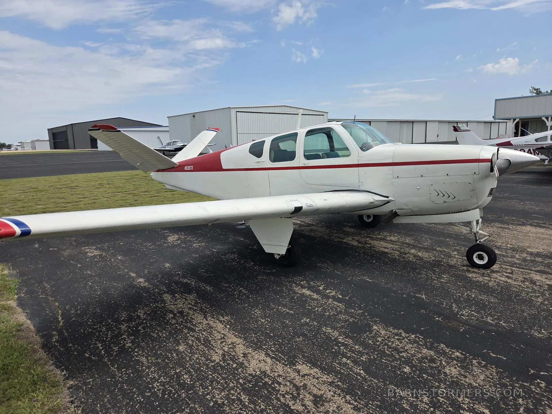 A white and red Beechcraft Bonanza aircraft with registration N535TX is parked on an asphalt ramp, viewed from the front right with hangars in the background.