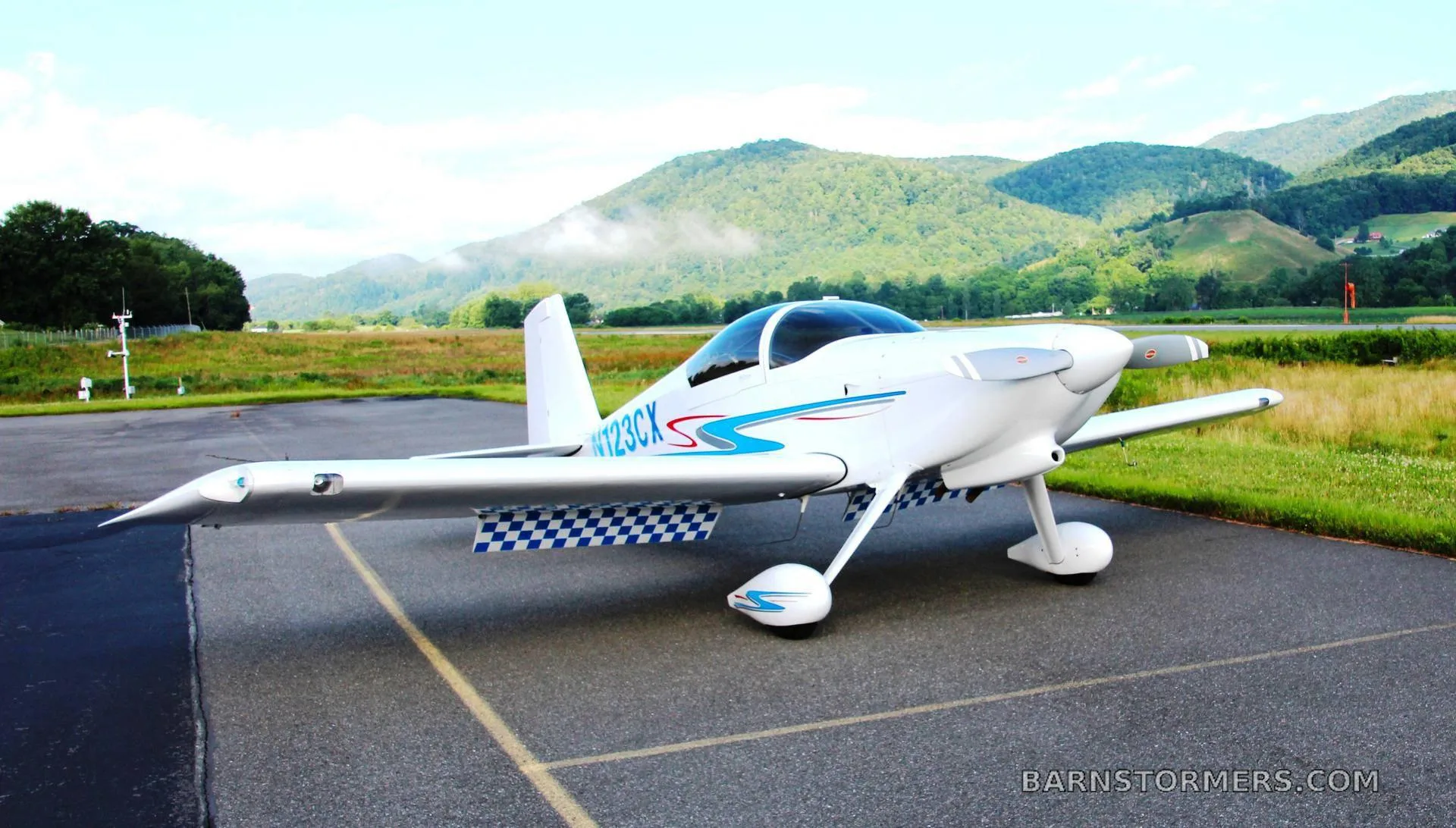A white single-engine aircraft with blue and red stripes (N123CX) parked on an airfield with green, misty mountains in the background.