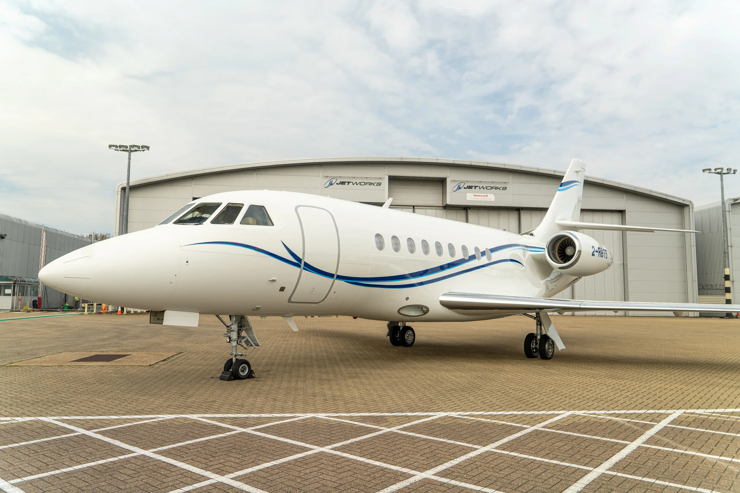 A white Dassault Falcon business jet with blue stripes is parked on an airfield in front of a hangar.