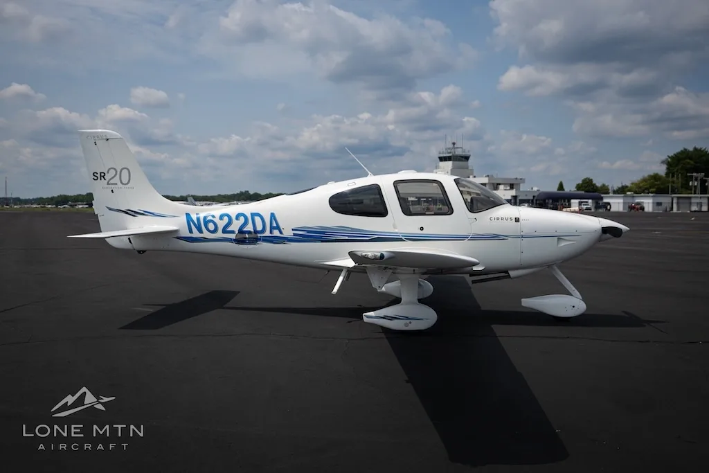A white Cirrus SR20 aircraft with blue accents and tail number N622DA parked on an airfield with a control tower in the background.