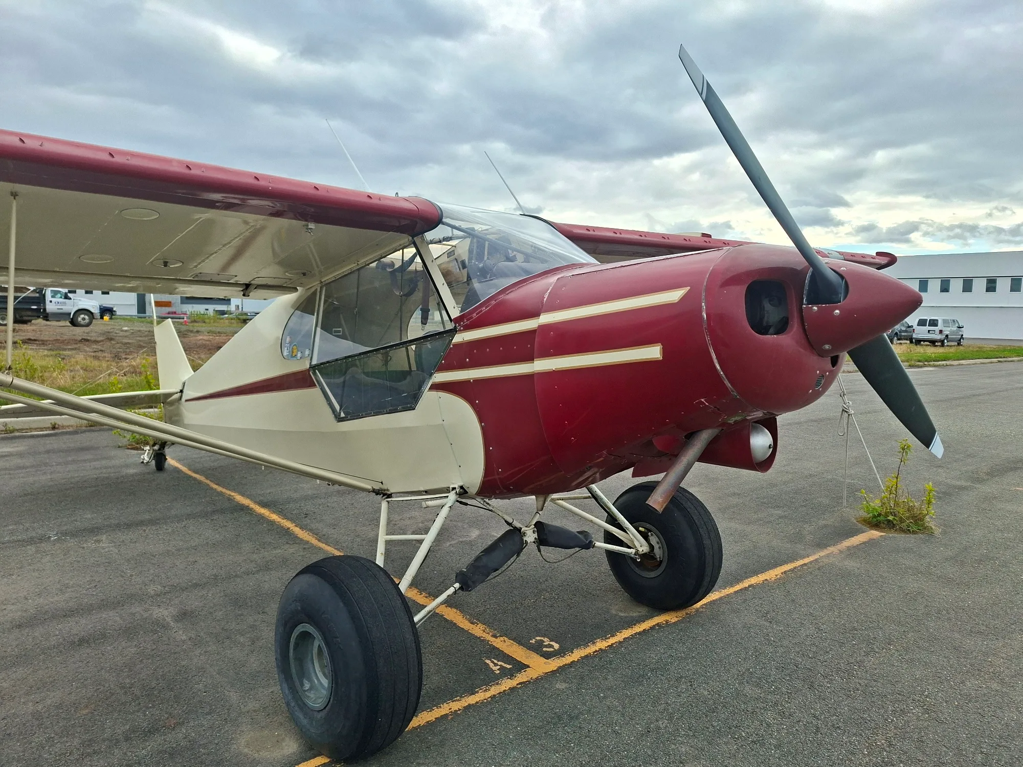 A red and cream high-wing bush plane with large tires, parked on an asphalt tarmac under a cloudy sky.