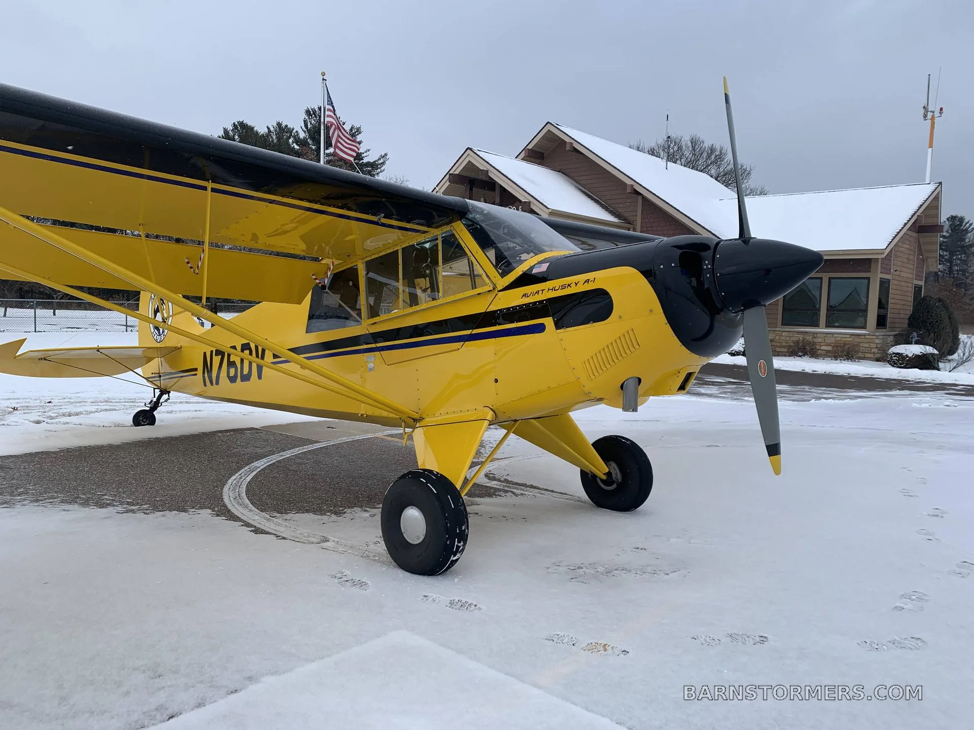 A yellow and black Aviat Husky A-1 aircraft with registration N76DV parked on a snowy airfield in front of a building.