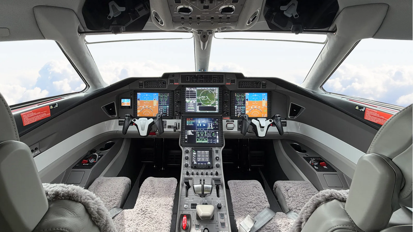 The modern glass cockpit of a business jet, featuring multiple large display screens and dual yokes, with clouds visible through the windshield.