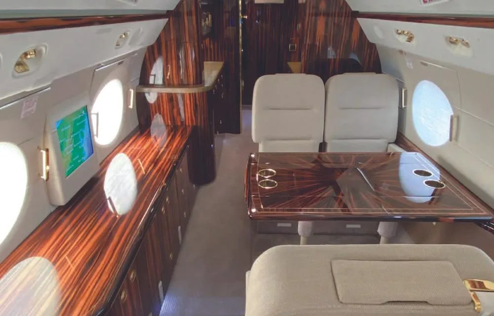 The private jet's cabin interior, featuring a dining or conference area with a polished wooden table and matching cabinetry.