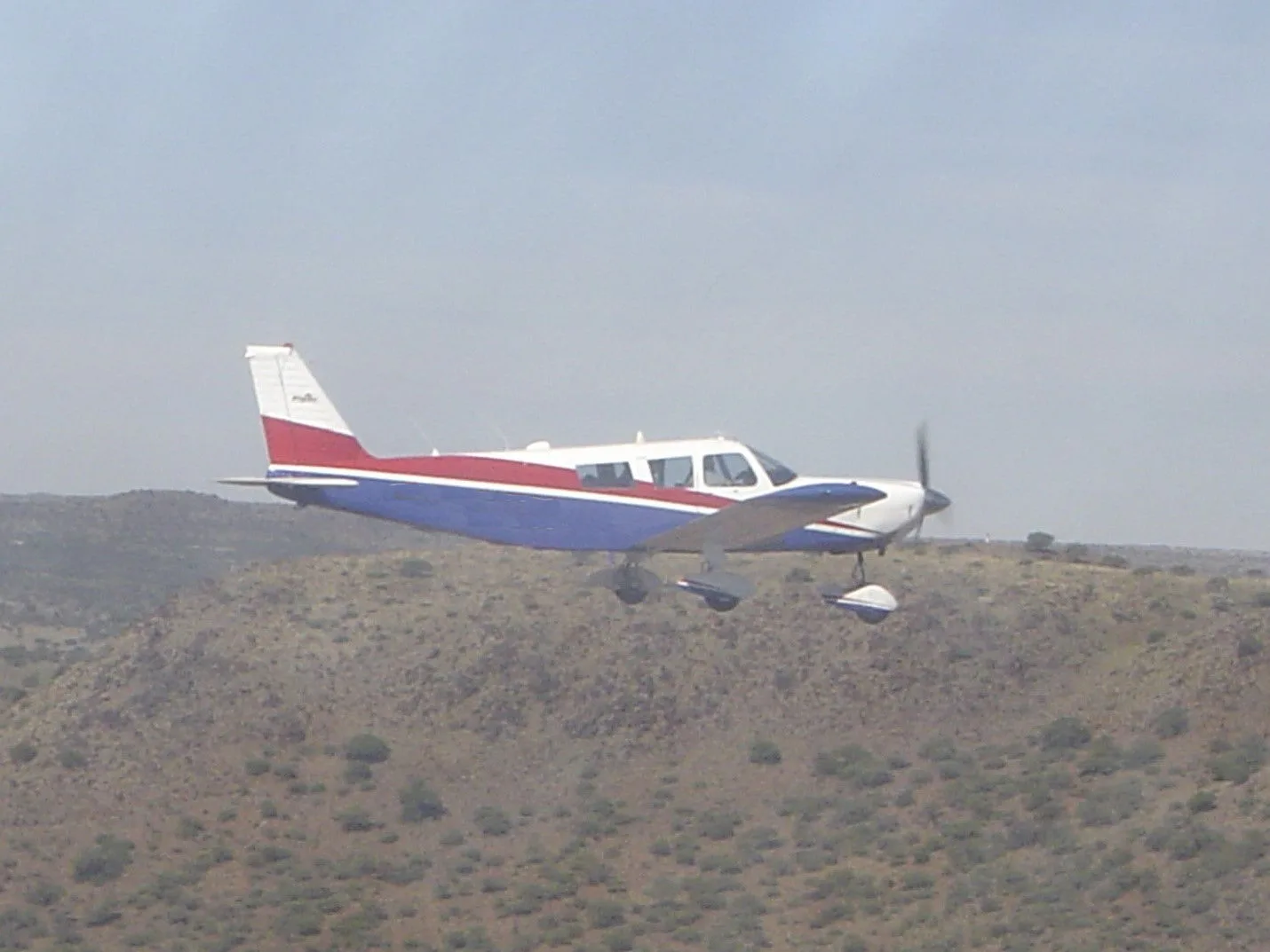 A white, red, and blue single-engine propeller aircraft flying over a dry, hilly landscape.