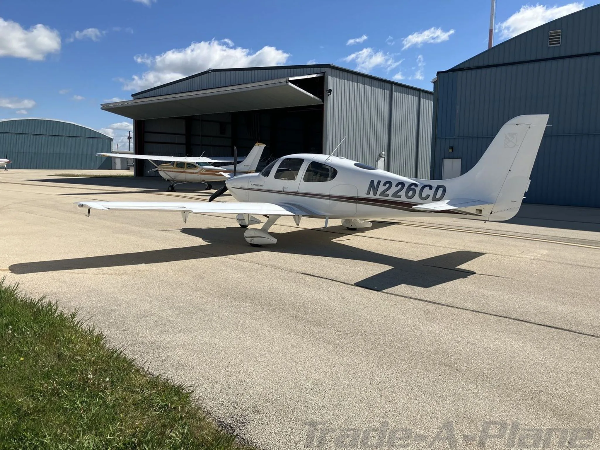 A white Cirrus SR20 aircraft with black and brown stripes, tail number N226CD, parked on an airfield tarmac with other aircraft and hangars in the background.