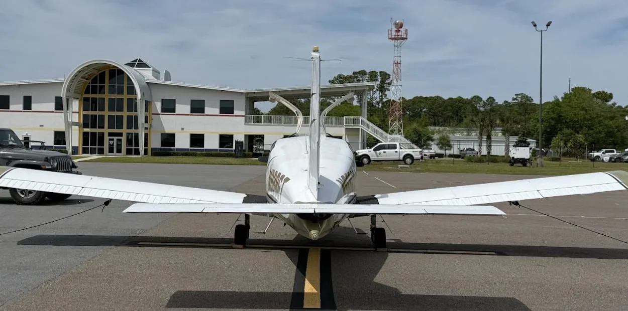 A rear view of the white and gold Socata TB-20 Trinidad aircraft with its doors open, parked in front of an airport building.