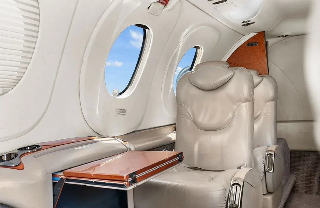A close-up of the private jet's beige leather cabin interior, showing an extended wooden side table and windows.