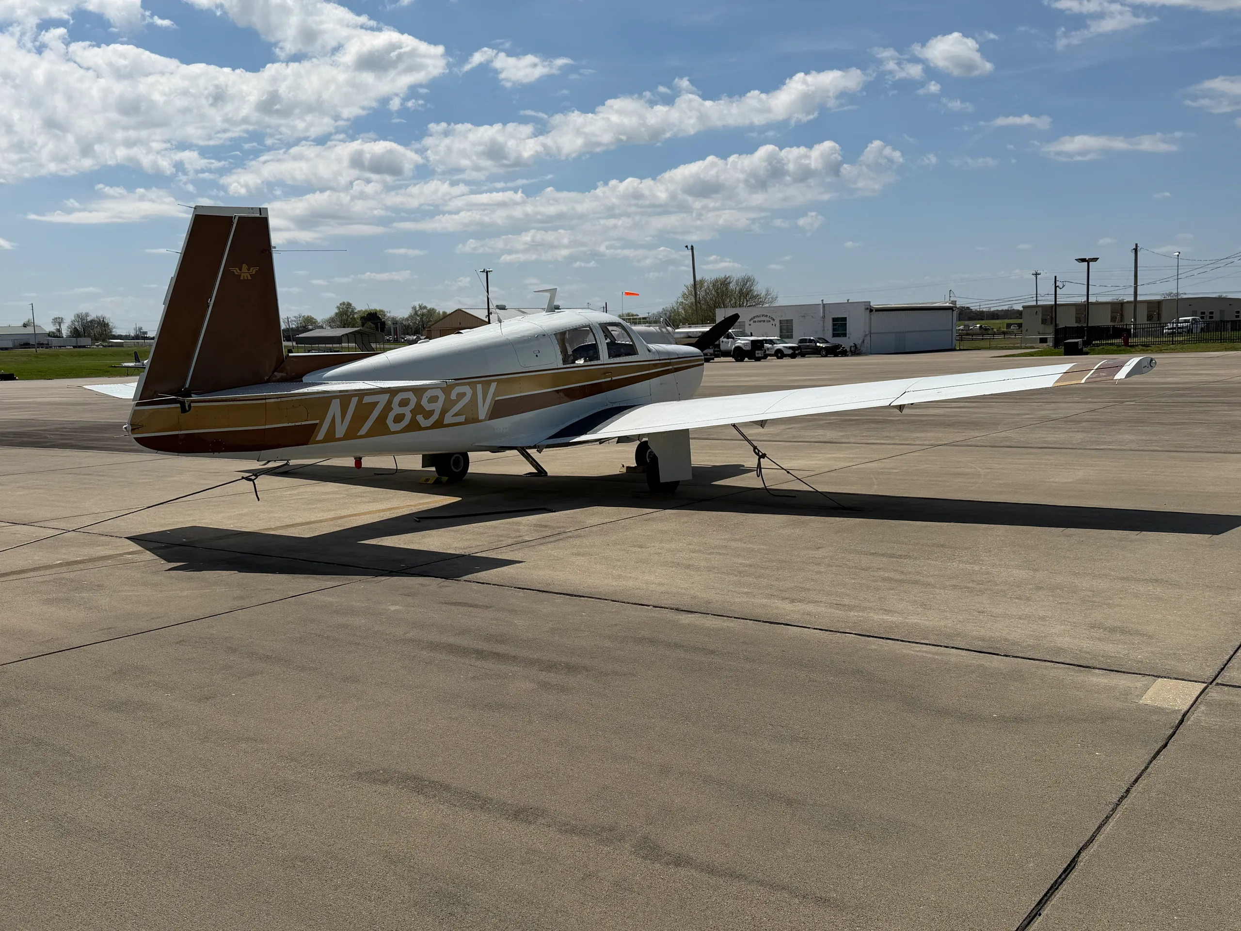 A white and brown single-engine aircraft (N7892V) parked on an airport tarmac with buildings in the background.