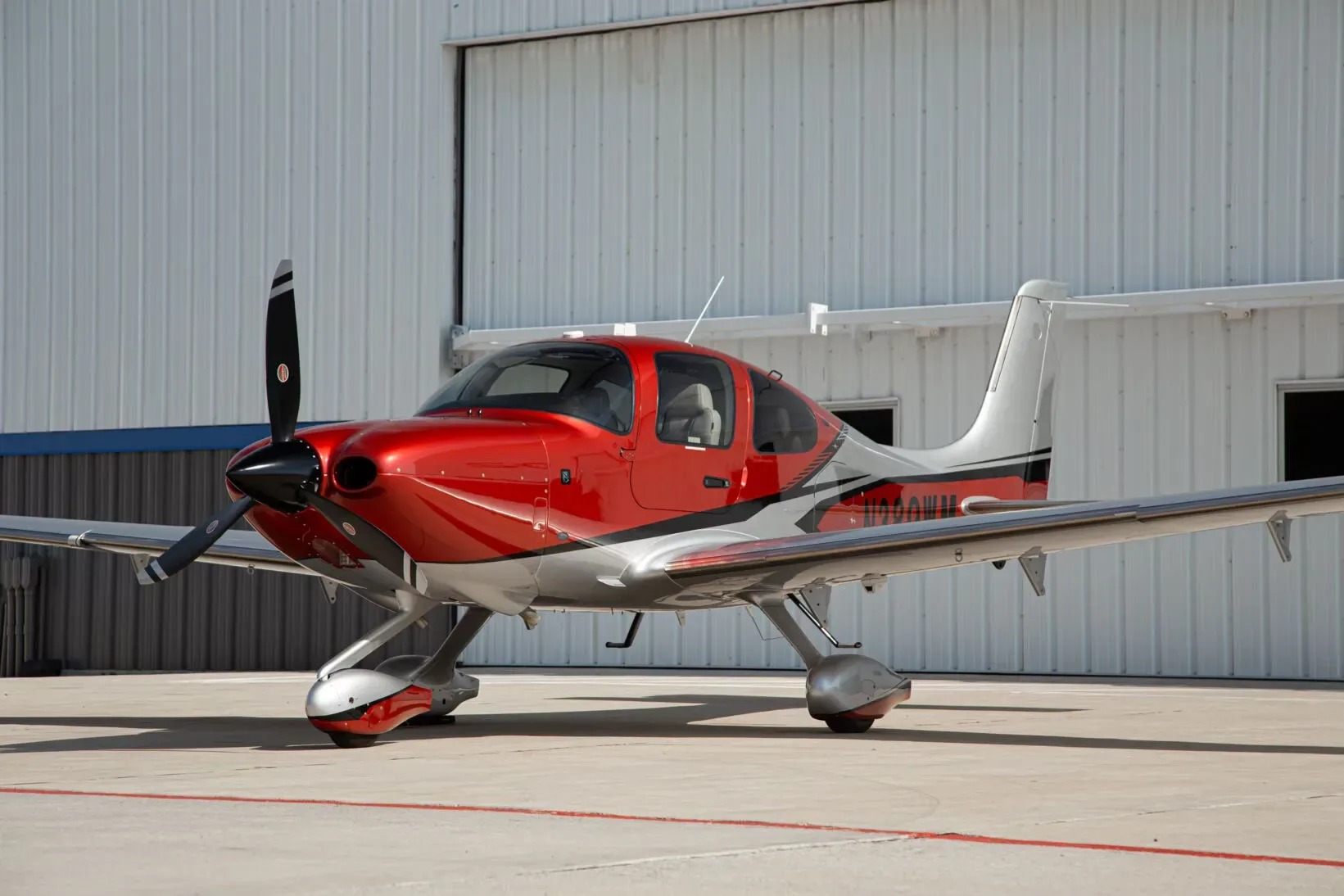 A red and silver single-engine propeller aircraft, N389WM, with its doors closed, parked on an airfield in front of a white hangar.
