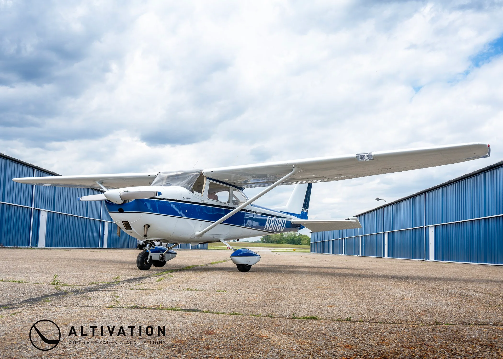 A white and blue Cessna 172 Skyhawk N8118U parked on an asphalt ramp between two blue hangars under a cloudy sky.