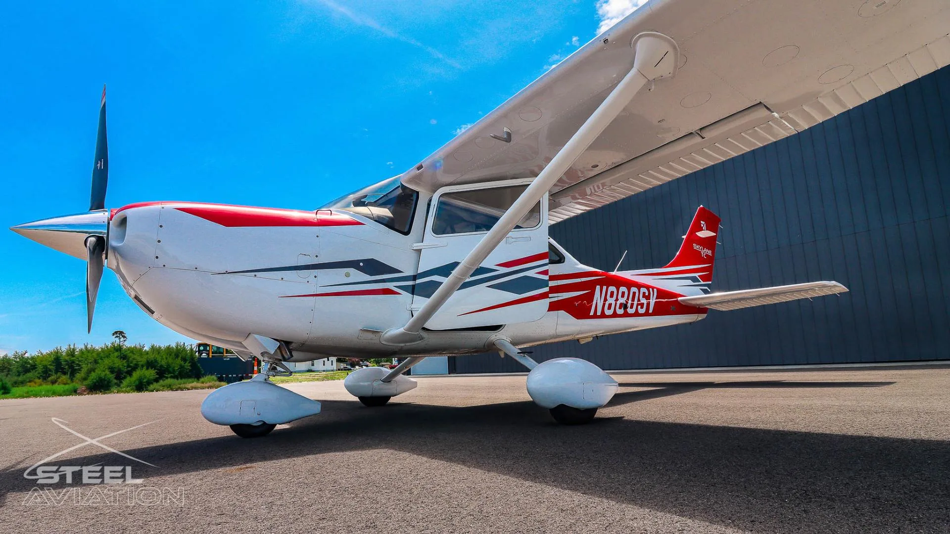 A white Cessna T182T Skylane TC with red and grey stripes is parked on an asphalt tarmac next to a dark hangar under a blue sky.