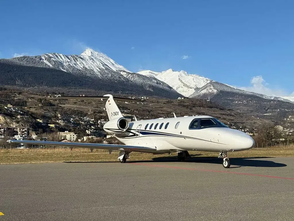 A white Cessna Citation jet with black and gold stripes is parked on an airfield with snow-capped mountains in the background.