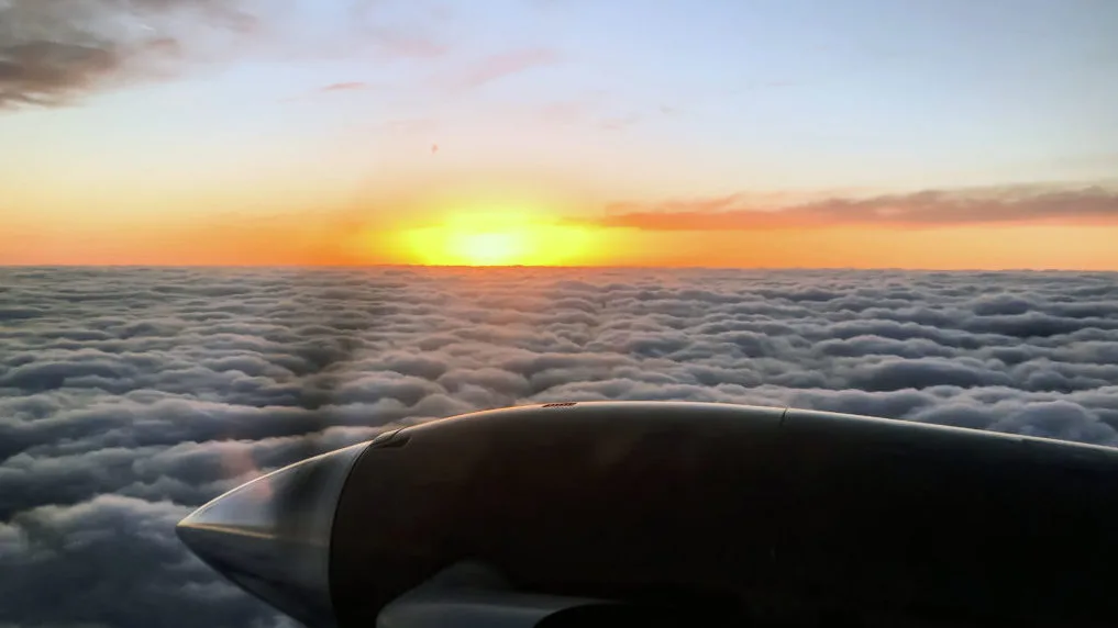 An aircraft engine nacelle is visible from a window, looking out over clouds at sunset.