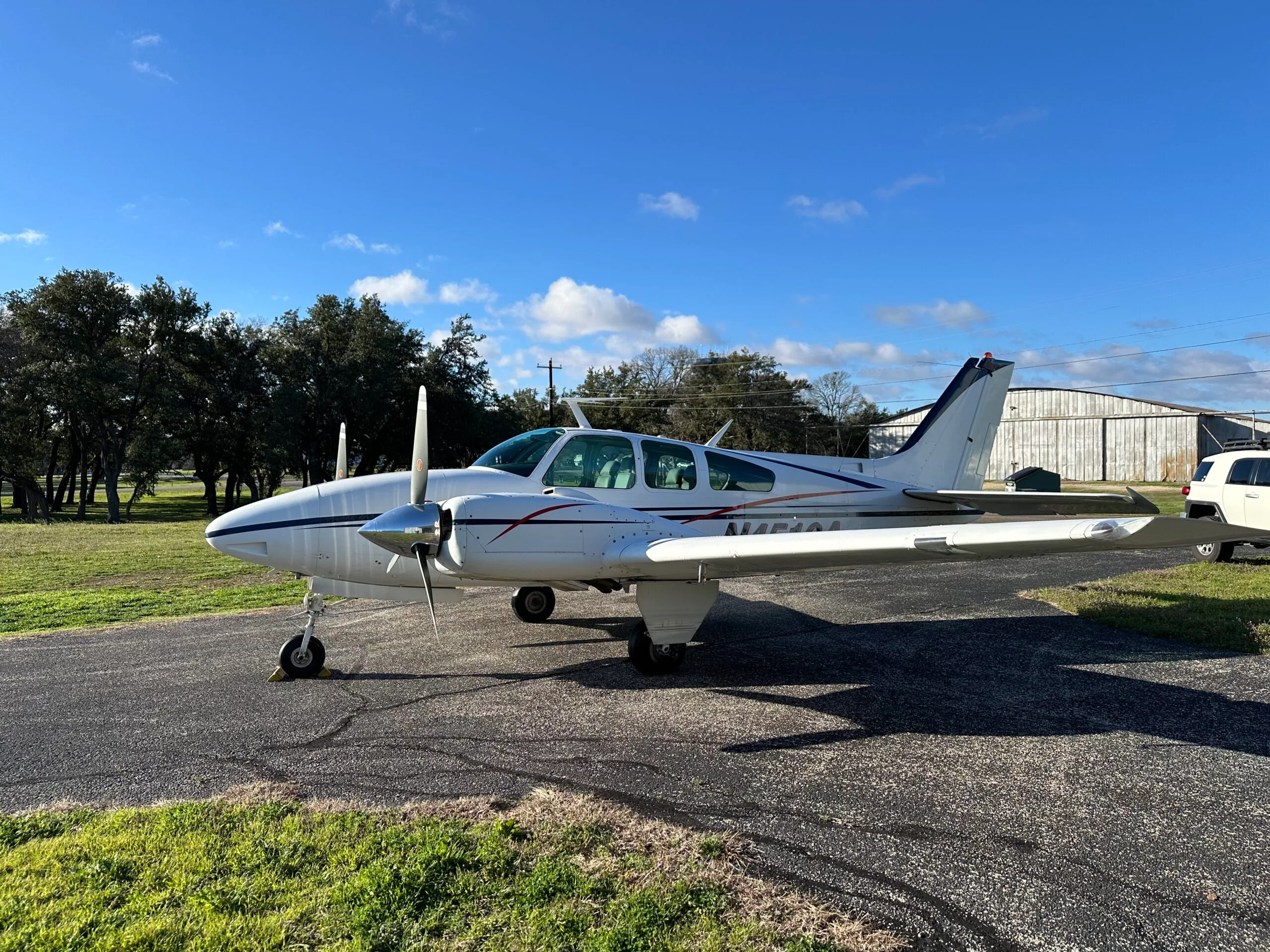 A white twin-engine propeller aircraft with blue and red stripes, registration N451CA, parked on an asphalt ramp with trees and a hangar in the background under a blue sky.
