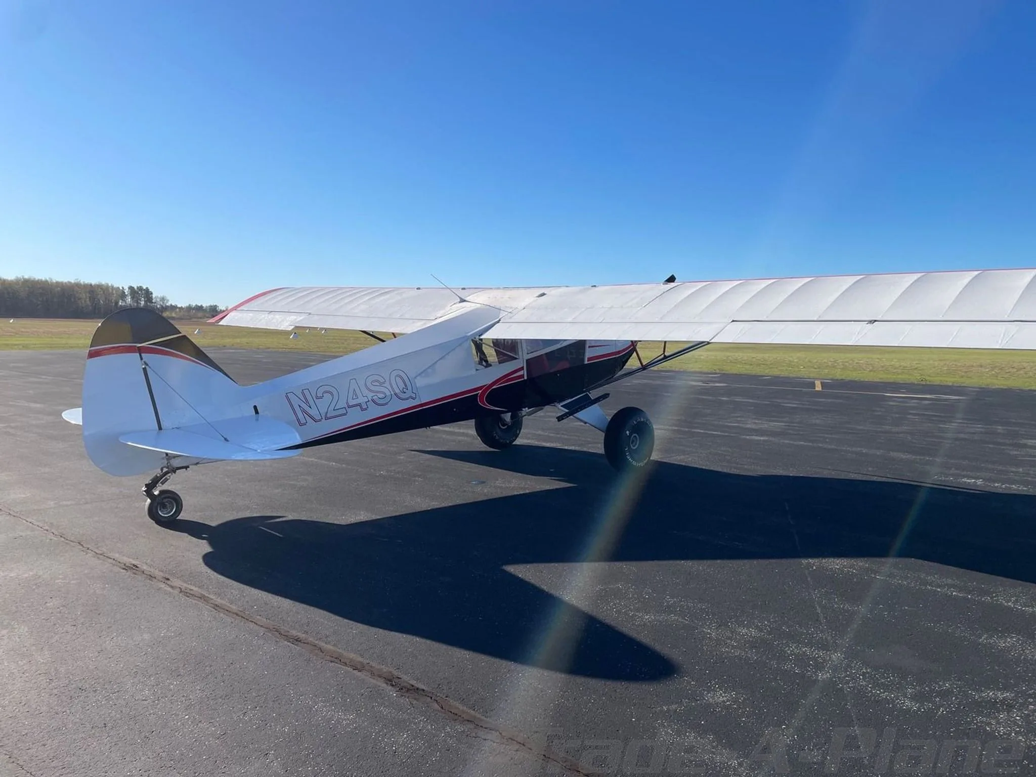 A rear-side view of the white, black, and red STOL Quest 4 aircraft (N24SQ) parked on an asphalt tarmac with a field in the background.