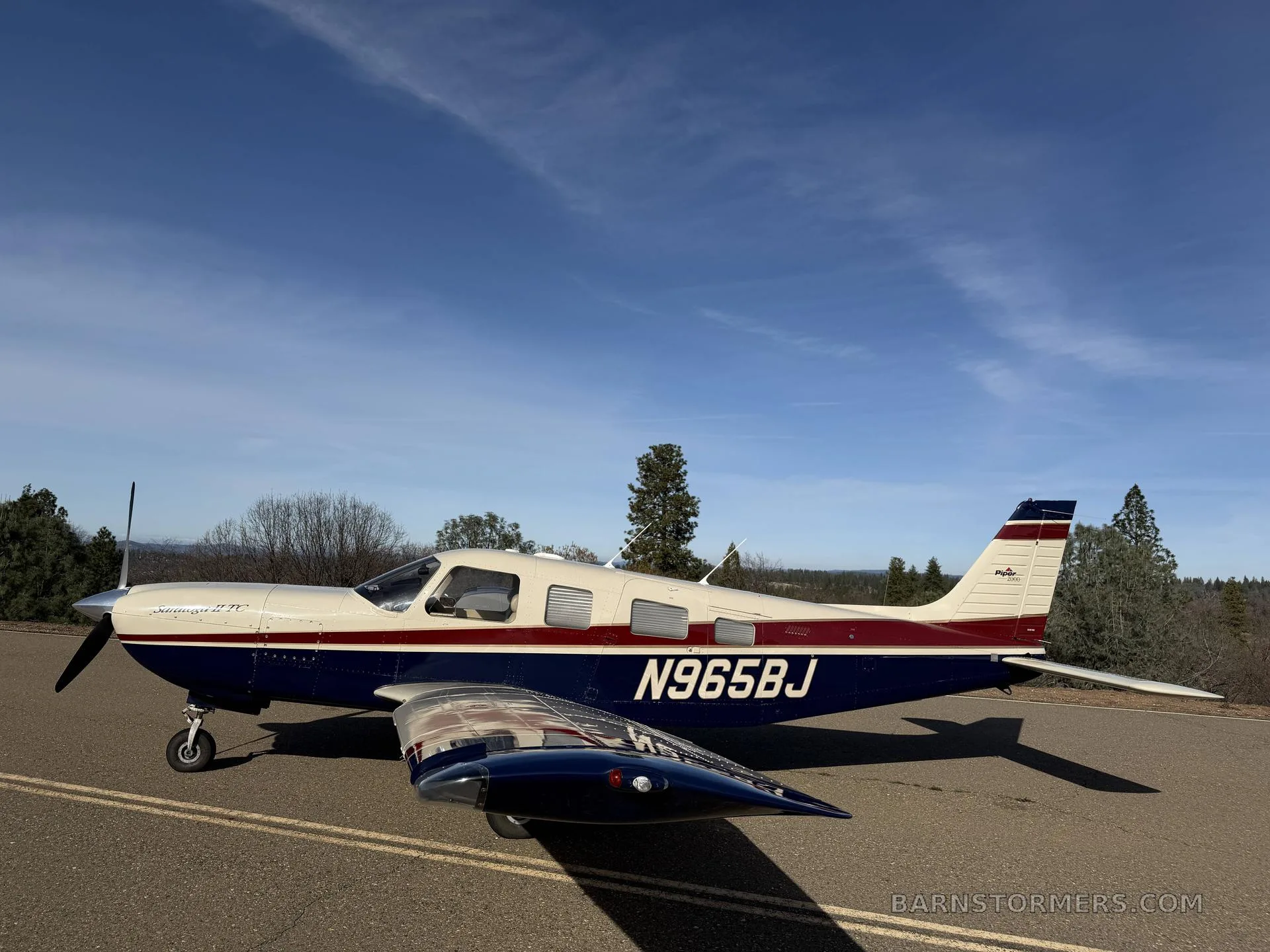 A side view of a white, red, and blue Piper Saratoga II TC aircraft (N965BJ) parked on an asphalt surface under a blue sky.