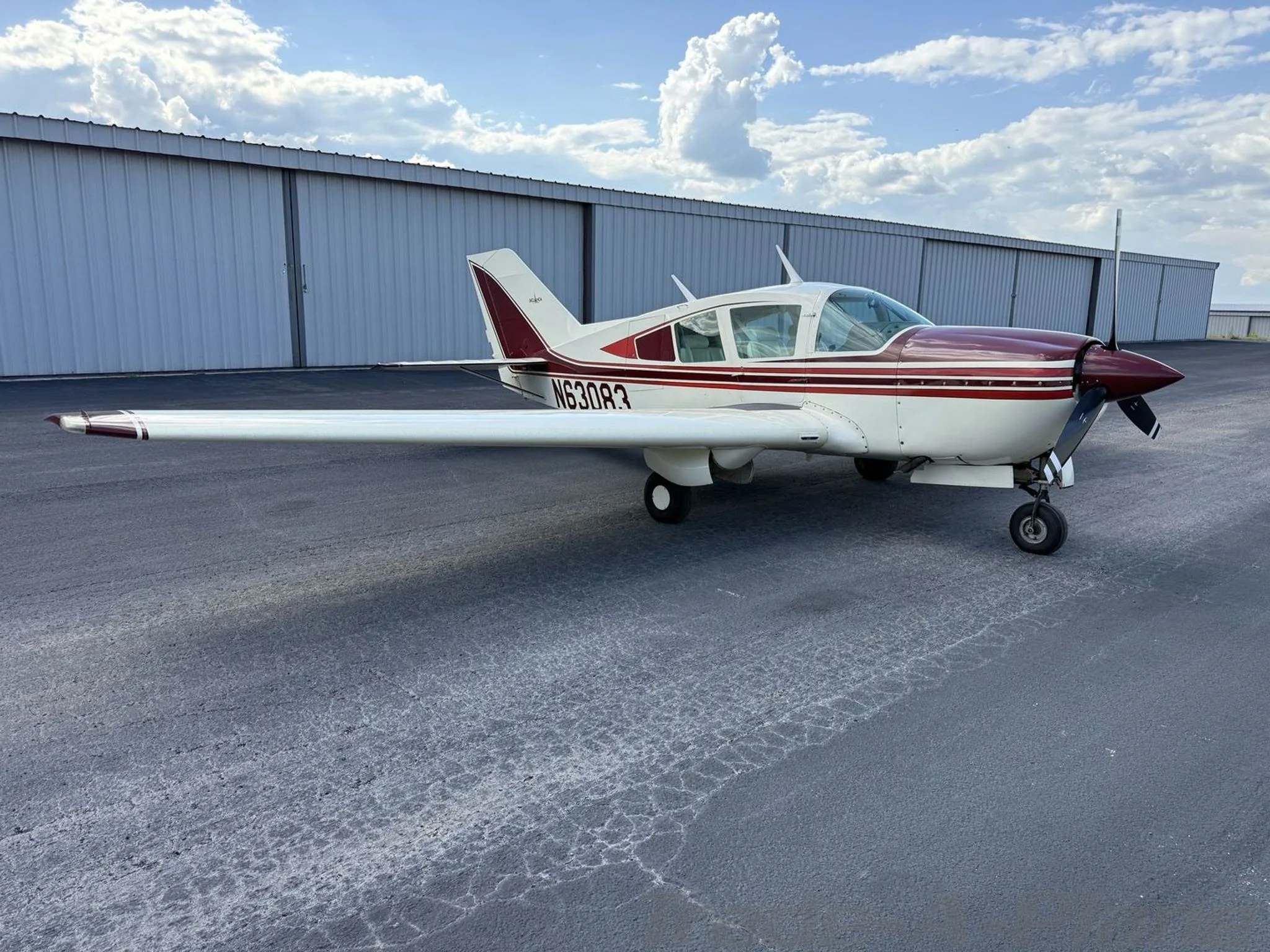 A white and maroon Bellanca Viking N63083 parked on a tarmac in front of hangars under a cloudy sky.