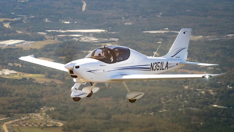 A white Tecnam P2008 light aircraft with blue stripes and registration N351LA is flying over a green, forested landscape with buildings.