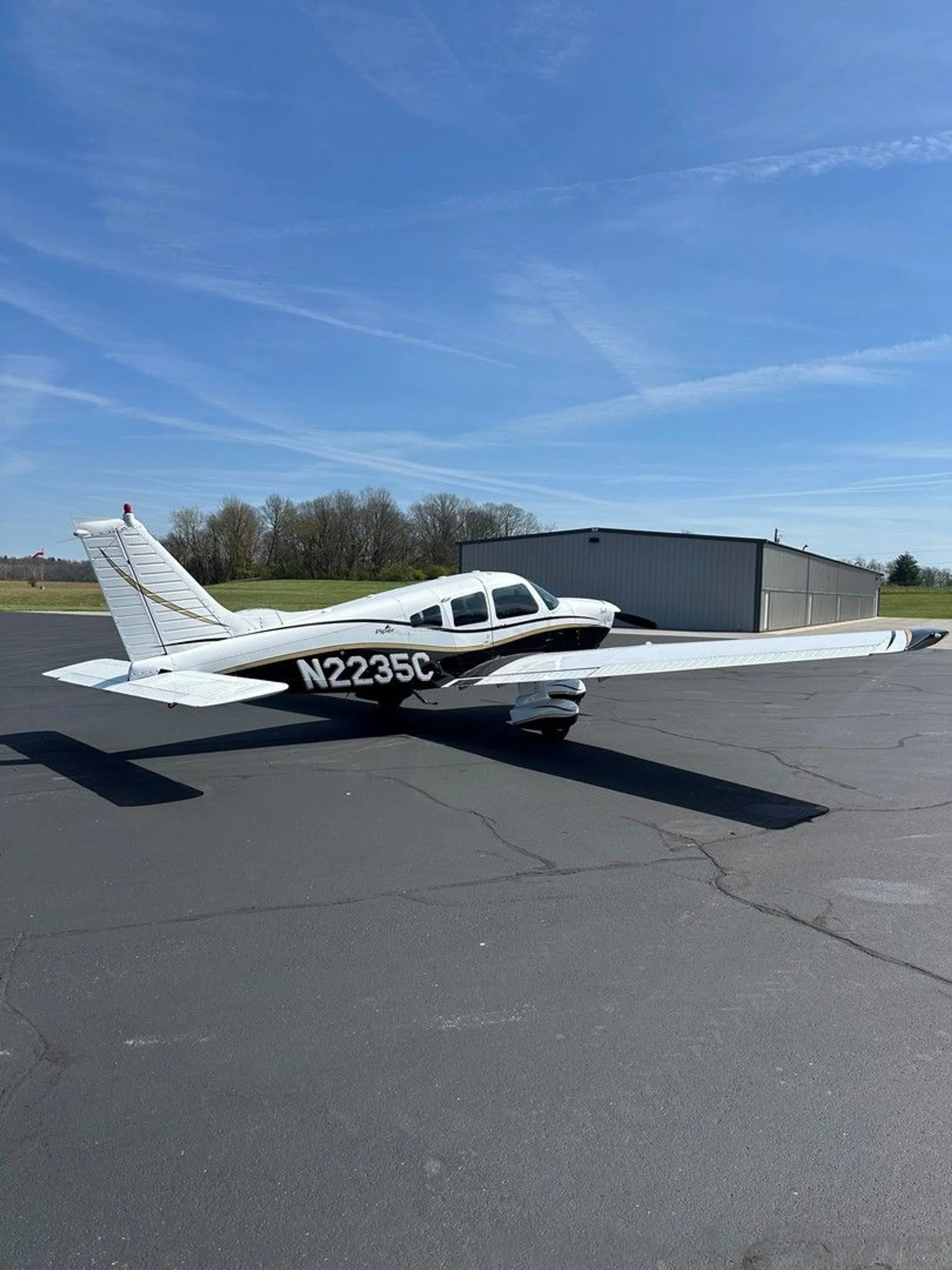 A white and black Piper aircraft with gold trim, registration N2235C, viewed from the rear-left on an asphalt ramp.