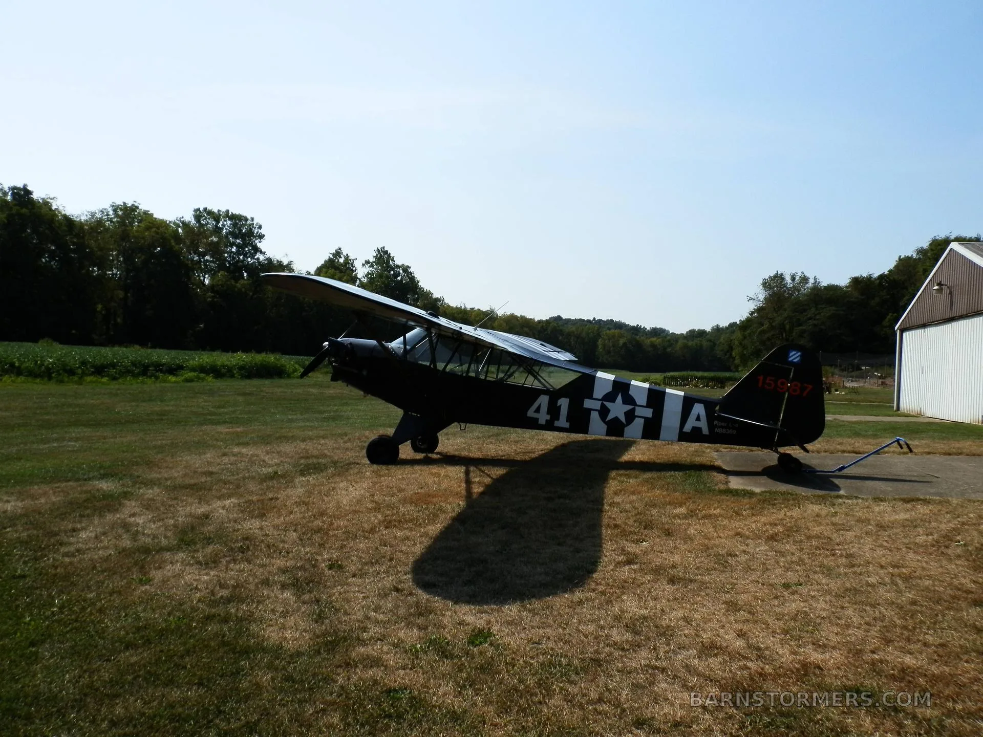 A black single-engine taildragger aircraft with white stripes and military-style markings parked on a grassy field next to a hangar.