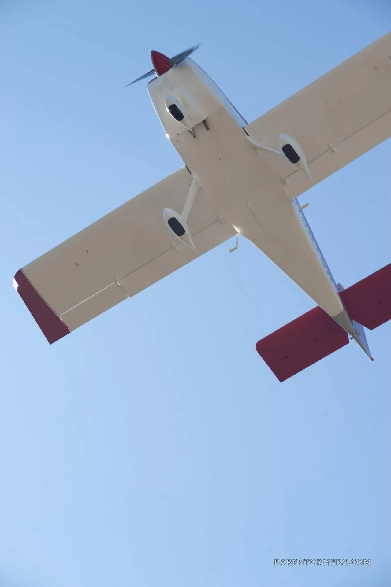 A red and white single-engine aircraft is seen from below, flying against a clear blue sky.