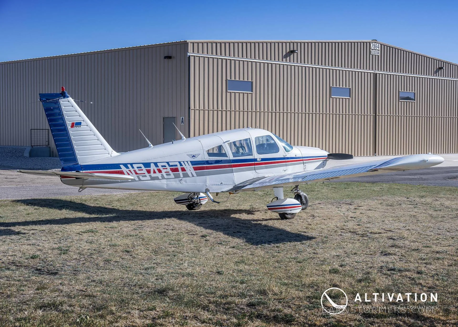 A white single-engine propeller aircraft with blue and red stripes is parked on grass next to a large beige hangar.