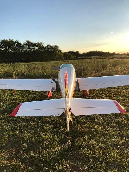 A rear view of a white, red, and yellow single-engine aircraft with a tail wheel on a grassy field at sunset.