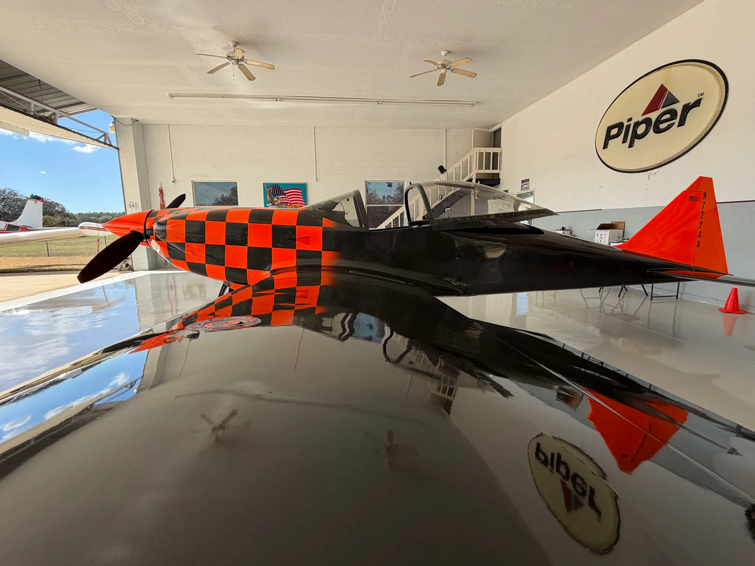 The black and orange checkered experimental aircraft, N7224A, is parked inside a hangar with a Piper logo on the wall.