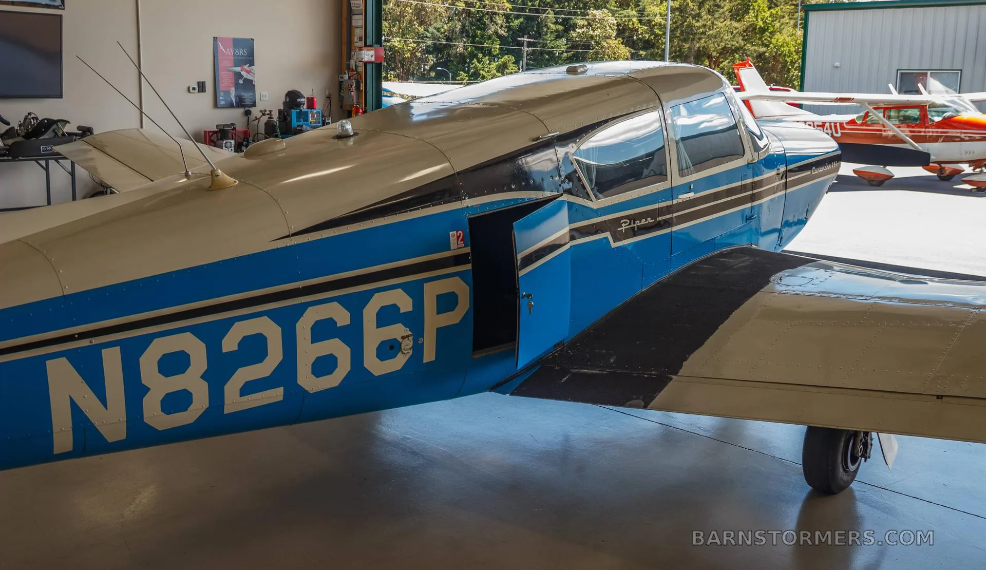 A side view of the blue and white Piper Comanche 250 (N8266P) with its cabin door open, parked in a hangar.