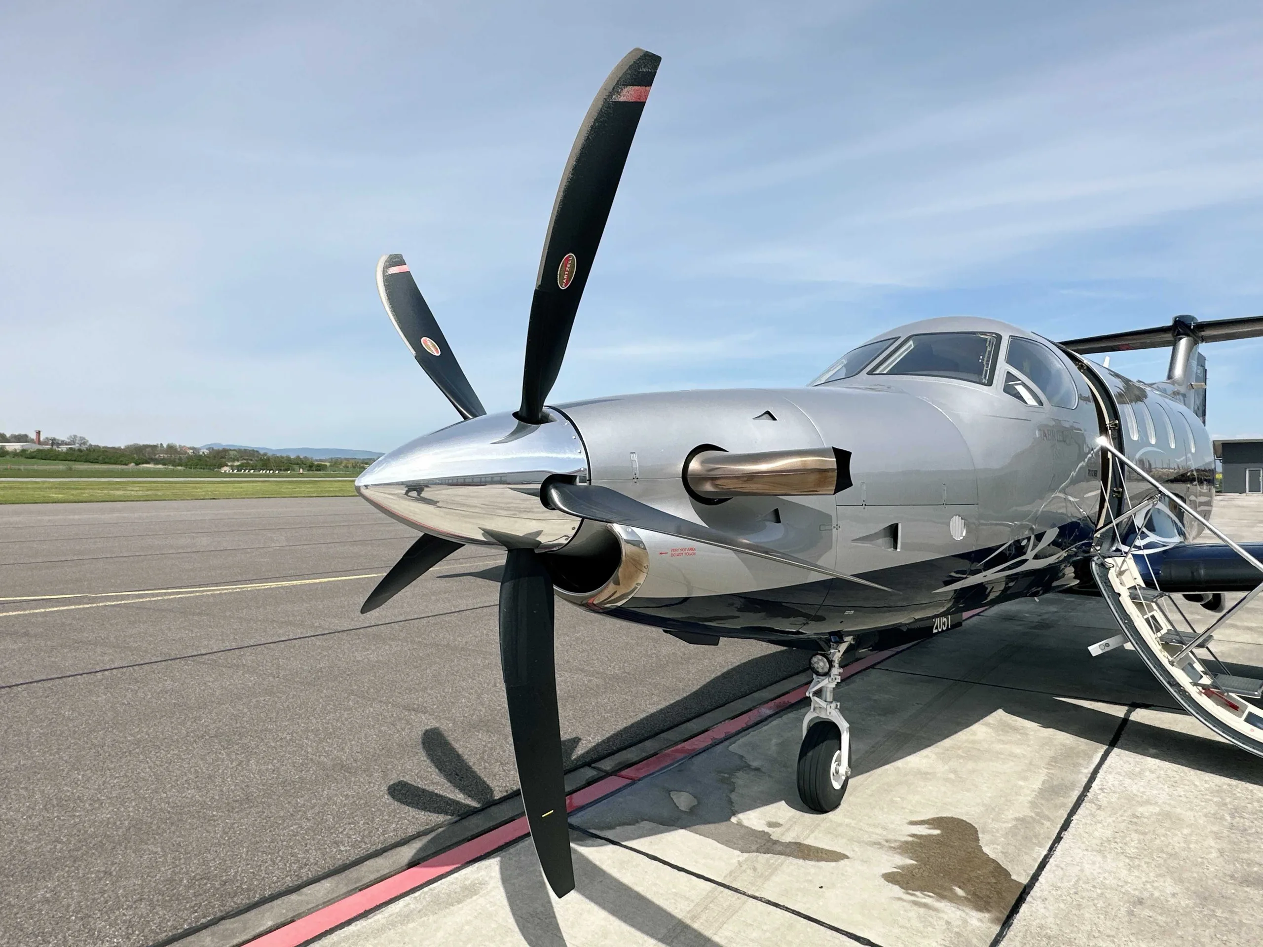 A close-up view of the nose and propeller of a silver and blue Pilatus PC-12 NGX aircraft on an airport tarmac.