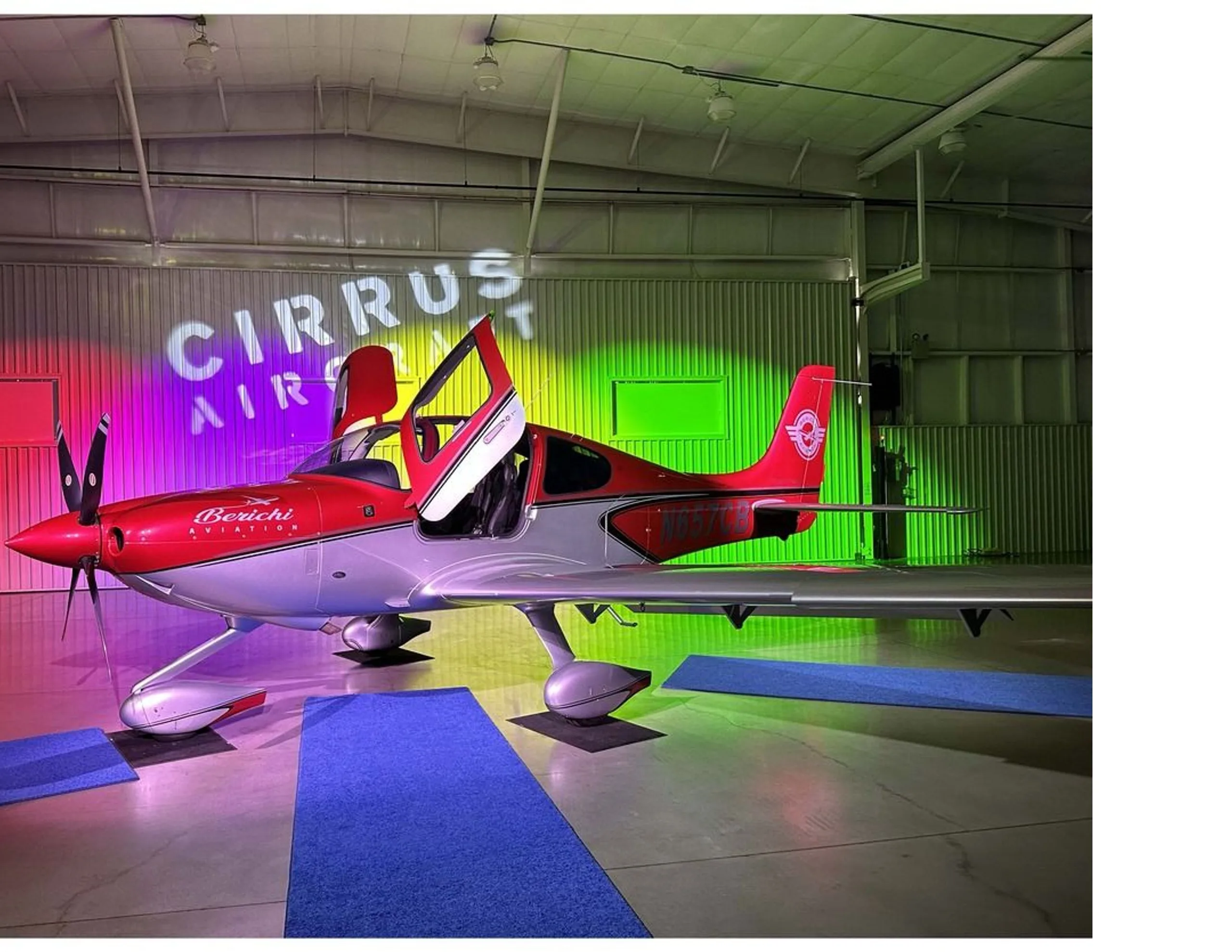 A wide side view of the red and silver Cirrus SR22 aircraft, N657CB, with open doors, in a hangar with "Cirrus Aircraft" projected on the wall.
