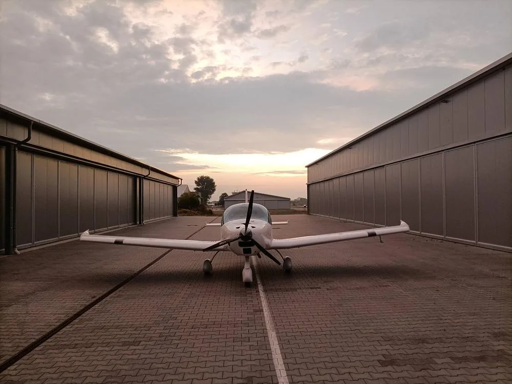 A white Bristell aircraft parked on a paved apron between two hangars at dusk.