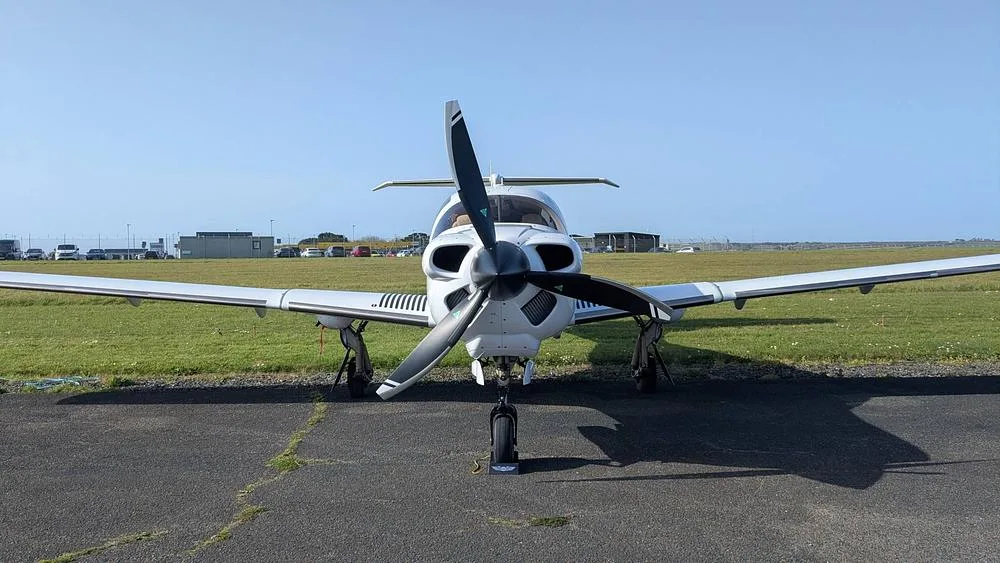 A white propeller aircraft with a black propeller, viewed head-on on an airfield under a clear sky.