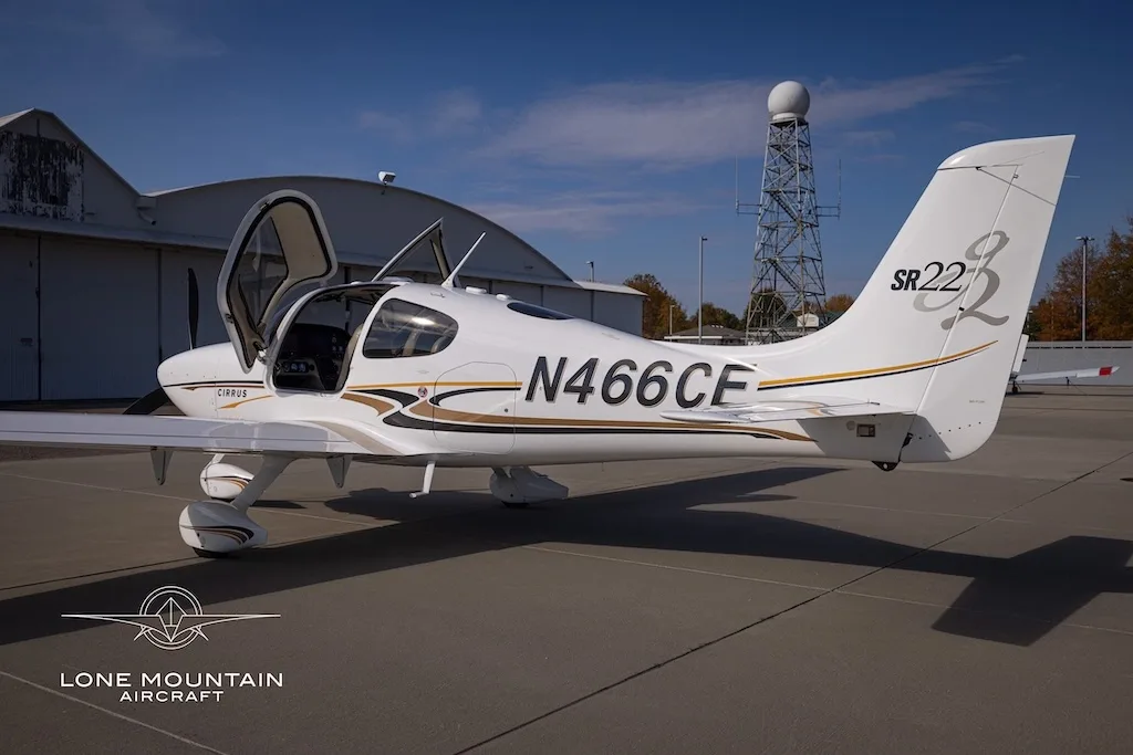 A white Cirrus SR22 aircraft with gold and black stripes, registration N466CE, parked on an airfield with its doors open, viewed from the rear-right.