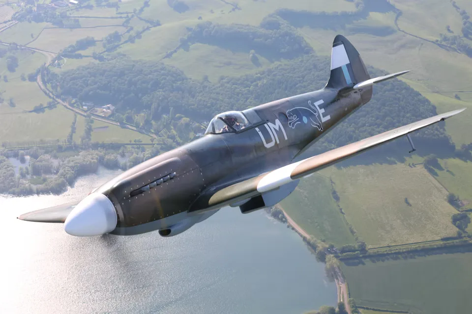 A Supermarine Spitfire in flight, banking over a lake and rolling green hills.