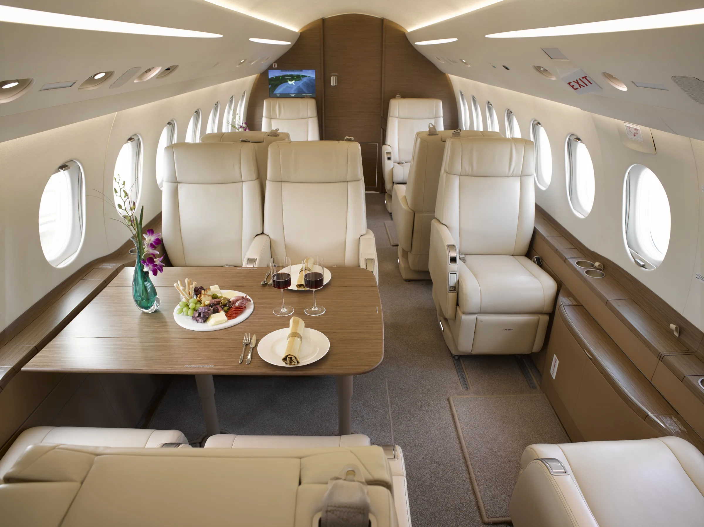 A spacious aircraft cabin interior featuring cream leather seating, a wooden dining table with a meal, and a monitor at the front.