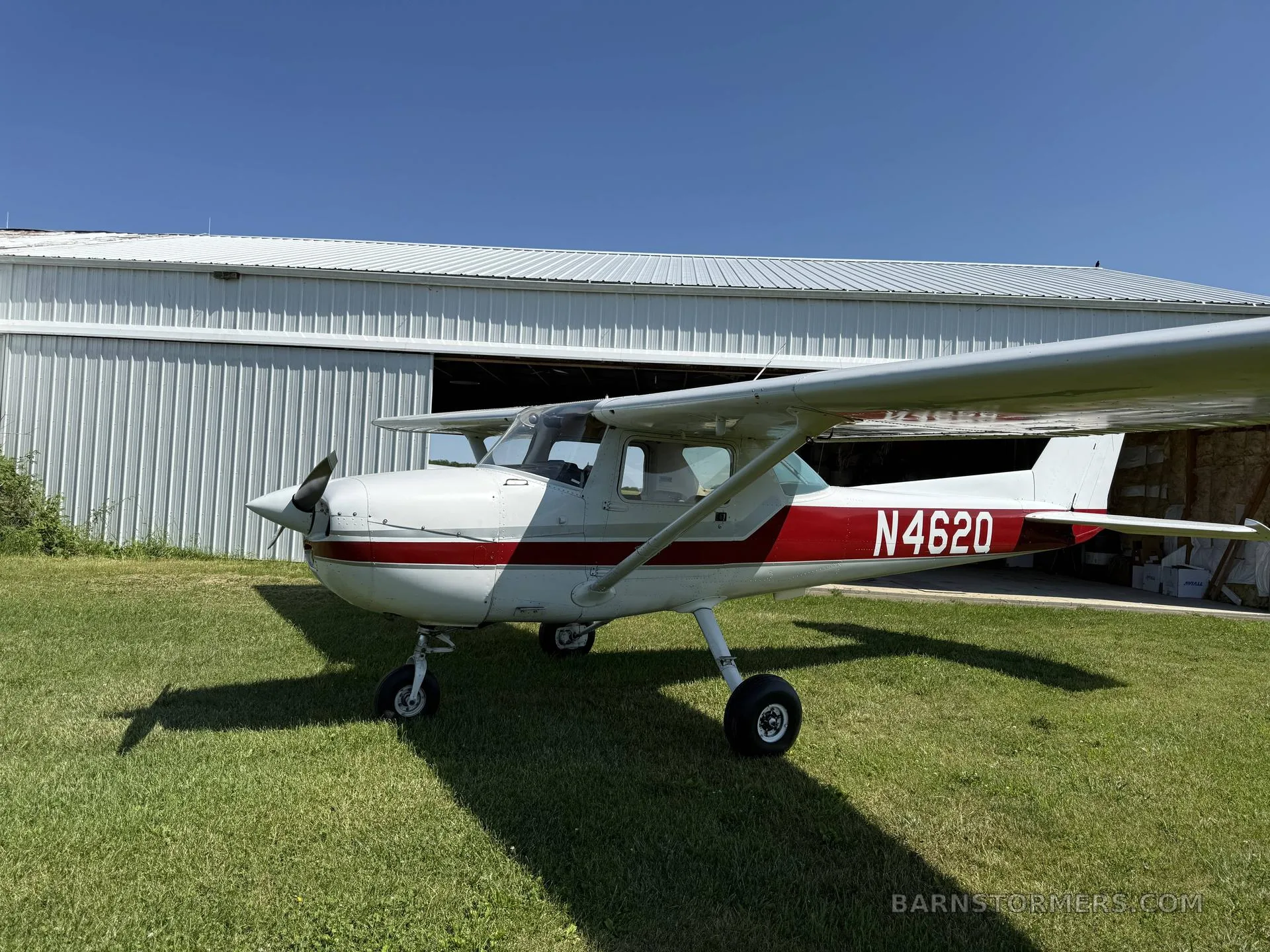 A white and red Cessna 150 is parked on a grassy field in front of a white hangar.