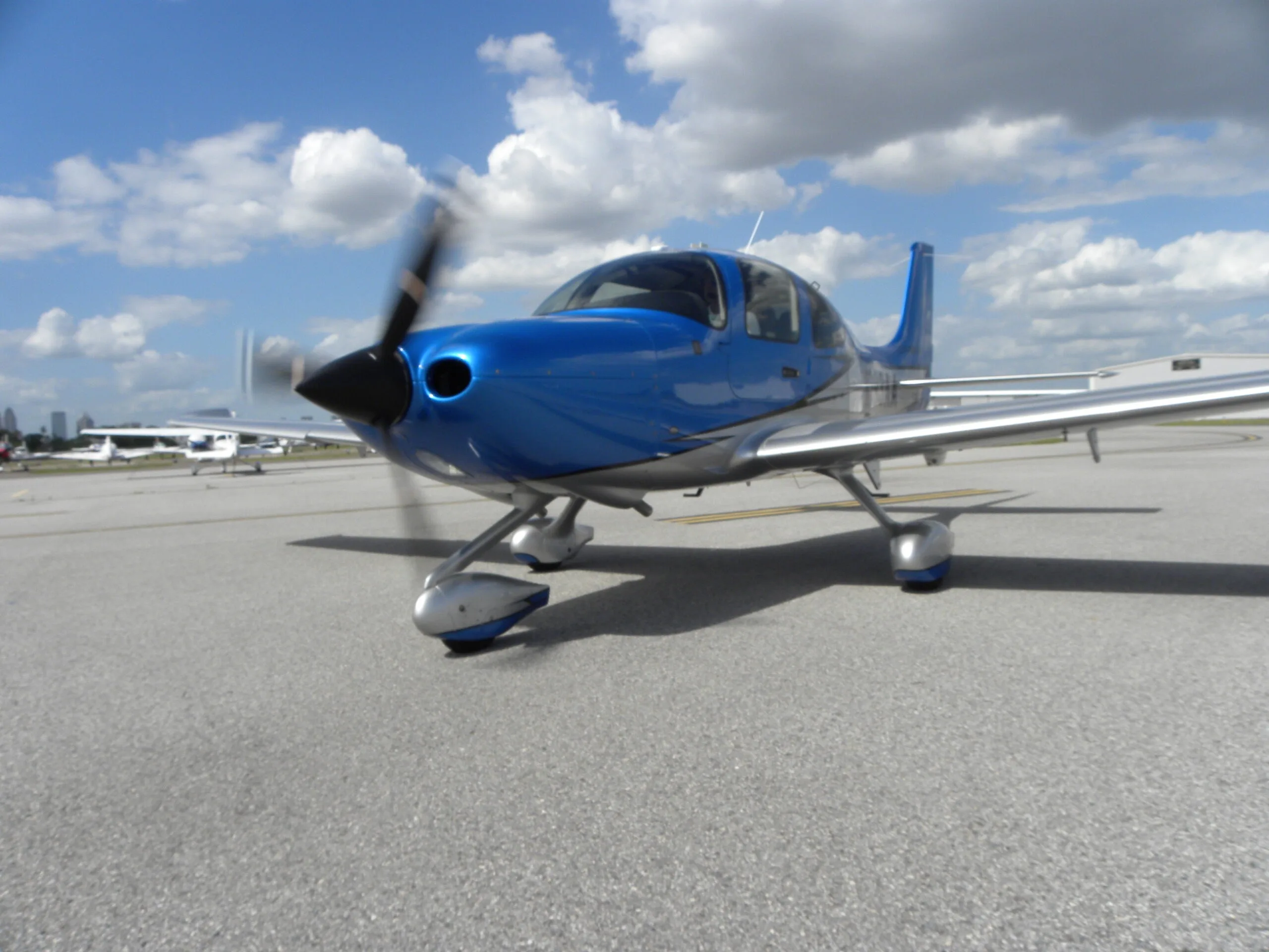 A blue and silver Cirrus SR22 aircraft with its propeller spinning, parked on an asphalt ramp.