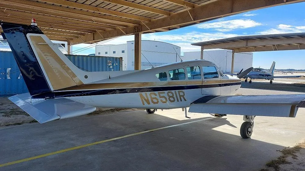 A white and gold single-engine propeller aircraft (Cessna 177 Cardinal) with registration N6581R is parked under an open-sided hangar, viewed from the rear-left.