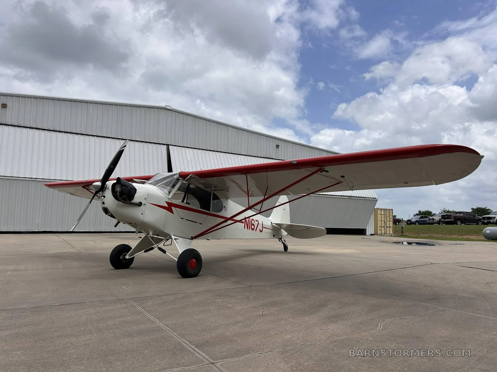 A white and red single-engine aircraft (N167J) with high wings is parked on an asphalt apron in front of hangars under a cloudy sky.
